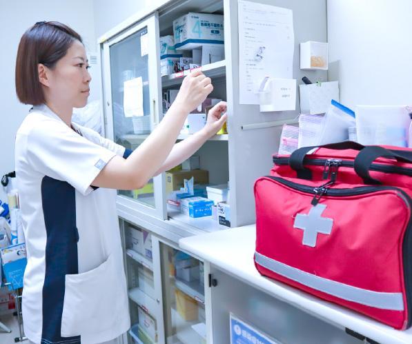 ナースステーションや薬品庫などの棚の前で、女性スタッフが医療資材や医薬品の確認・管理を行っている様子(手前に赤い救急バッグ)