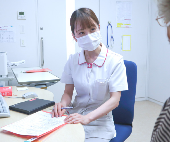 診察室(または相談室)にて、マスクを着用した白いユニフォームの女性スタッフが机を挟んで患者(または家族)の相談に熱心に耳を傾けている様子