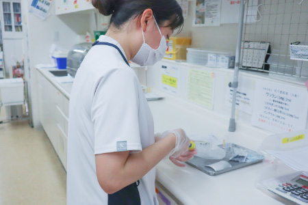 手袋とマスクを着用した白いユニフォームの女性スタッフが、作業台で薬品や医療器具の準備を行っている様子