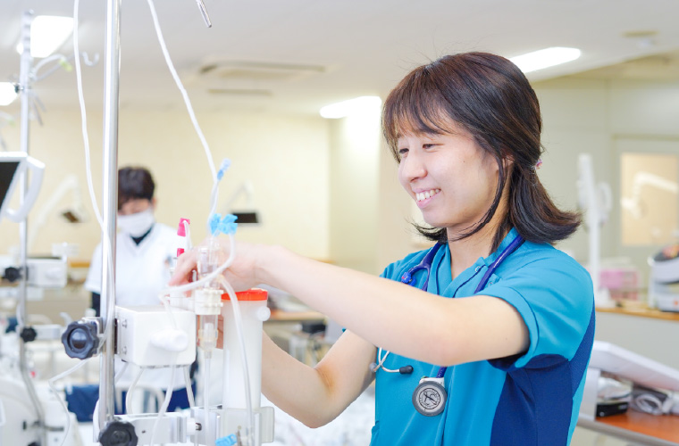 鮮やかなブルーのスクラブを着用した女性スタッフが、笑顔で透析機器の操作や確認を行っている様子